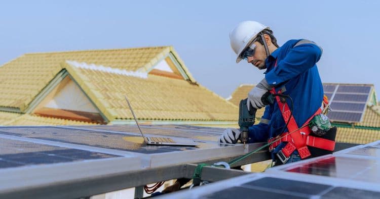 Ein Monteur in einem Kletter-Harnisch, der auf einem Dach in einer Wohnsiedlung eine Solaranlage installiert.